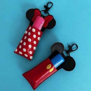 May include: Two red and black lip balm holders with keychains. One holder features a red and white polka dot pattern and a pink lip balm. The other holder features a black Mickey Mouse design and a blue lip balm.