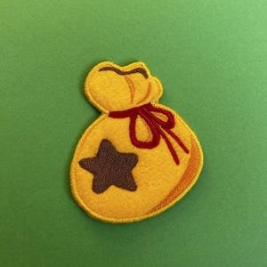 May include: A yellow felt patch of a money bag with a red ribbon and a brown star. The bag is embroidered with a yellow outline.
