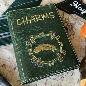 May include: A green leather-bound book with the title "CHARMS" embossed in gold lettering. The cover features a gold embroidered feather inside a decorative circle with purple accents.