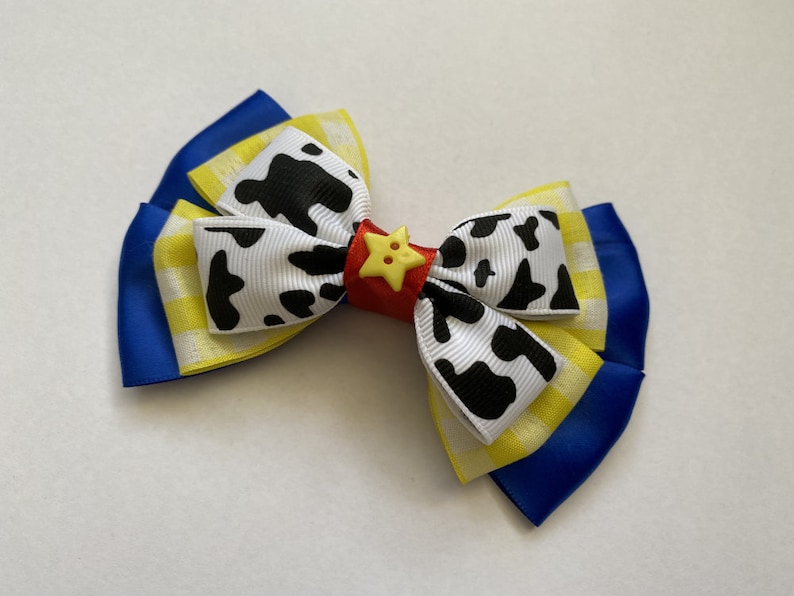 Pu&ograve; includere: Un fiocco blu, giallo e bianco con stampa di mucca e un bottone a stella rosso e giallo.