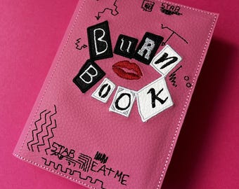 以下が含まれることがあります： ピンクの革製のノートブックで、前面に「Burn Book」という文字が黒と白の文字で刺繍されています。文字は赤い口紅のキスを囲むように円形に配置されています。ノートブックには、黒で「Stab Eat Me」という文字も刺繍されています。