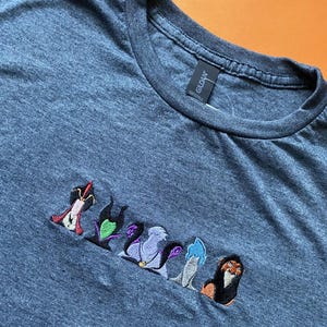 Könnte beinhalten: Ein dunkelgraues T-Shirt mit gestickten Bildern von Disney-Bösewichten. Zu den gestickten Figuren gehören Maleficent, Ursula und Scar. Das Shirt besteht aus einem weichen Material und hat einen Rundhalsausschnitt.