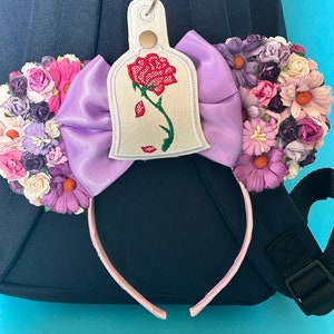 K&ouml;nnte beinhalten: Ein dekoratives Stirnband mit einer gro&szlig;en lavendelfarbenen Schleife und floralen Akzenten. Das Stirnband enth&auml;lt einen Anh&auml;nger mit einem gestickten roten Rosenmuster. Das Accessoire befindet sich vor einem blauen Hintergrund und einem dunkelblauen Rucksack.