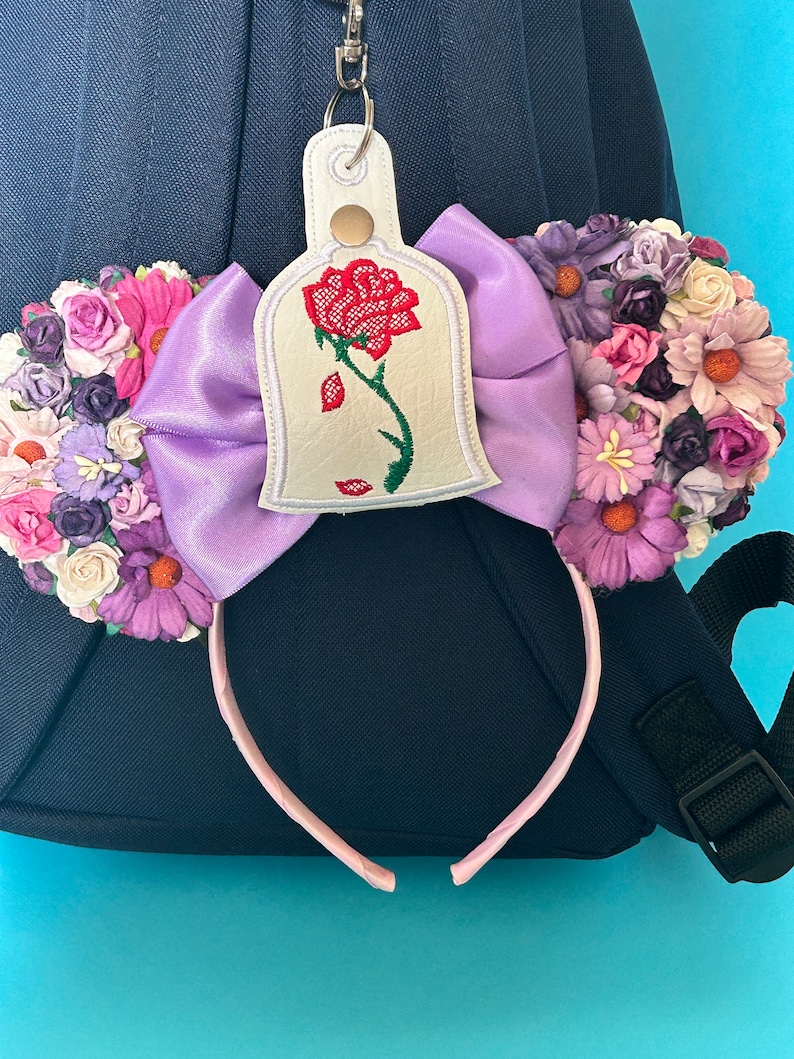 K&ouml;nnte beinhalten: Ein dekoratives Stirnband mit einer gro&szlig;en lavendelfarbenen Schleife und floralen Akzenten. Das Stirnband enth&auml;lt einen Anh&auml;nger mit einem gestickten roten Rosenmuster. Das Accessoire befindet sich vor einem blauen Hintergrund und einem dunkelblauen Rucksack.