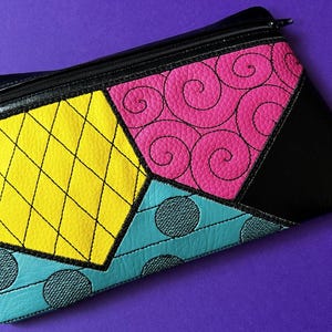 May include: A black zippered pouch with a patchwork design. The patches are yellow with black stitching, pink with black swirls, turquoise with black dots, and black.