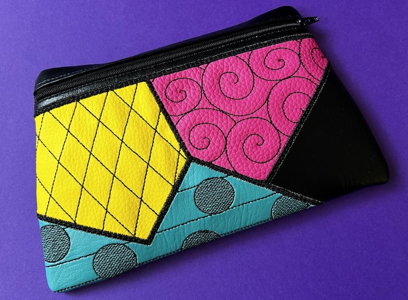 May include: A black zippered pouch with a patchwork design. The patches are yellow with black stitching, pink with black swirls, turquoise with black dots, and black.