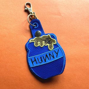 May include: Blue leather keychain shaped like a honey pot with a gold honey design and the word "Hunny" on the front.