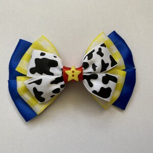 Pu&ograve; includere: Un fiocco blu, giallo e bianco con un motivo a stampa di mucca bianco e nero e un bottone a stella giallo al centro.