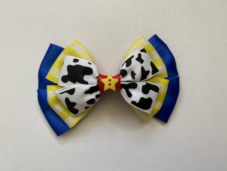 Pu&ograve; includere: Un fiocco blu, giallo e bianco con un motivo a stampa di mucca bianco e nero e un bottone a stella giallo al centro.