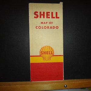 Peut inclure: Une carte routière vintage de Shell pour le Colorado. La carte est imprimée sur du papier crème avec une bordure jaune et rouge. Le logo Shell est une coquille jaune avec le mot "Shell" en lettres rouges.