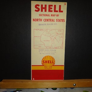 Peut inclure: Une carte routière vintage Shell des États du centre-nord, avec une carte du Minnesota, du Wisconsin et du Michigan. La carte est imprimée sur du papier crème avec du texte rouge et noir. La carte est bordée d'un cadre rouge avec le logo Shell au centre en bas. Le texte "Norb's Shell Service" est imprimé sous la carte avec l'adresse et le numéro de téléphone.