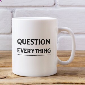 May include: White ceramic coffee mug with black text that reads "QUESTION EVERYTHING" on a wooden table with a vase of purple flowers.