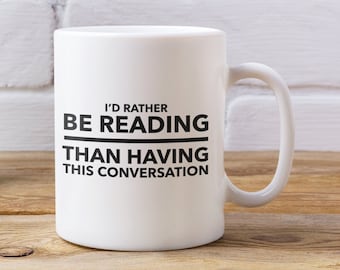 May include: A white ceramic mug with the text "I'D RATHER BE READING THAN HAVING THIS CONVERSATION" in black. A turquoise pitcher with white polka dots holds purple flowers. The items are on a wooden surface.