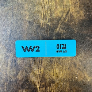 以下が含まれることがあります： 左側に「WW2」の文字、右側に韓国語で「이걸」と「새기는 순간」と書かれた青い長方形のネームタグです。