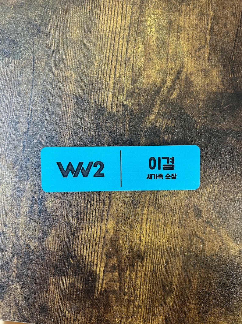 以下が含まれることがあります： 左側に「WW2」の文字、右側に韓国語で「이걸」と「새기는 순간」と書かれた青い長方形のネームタグです。