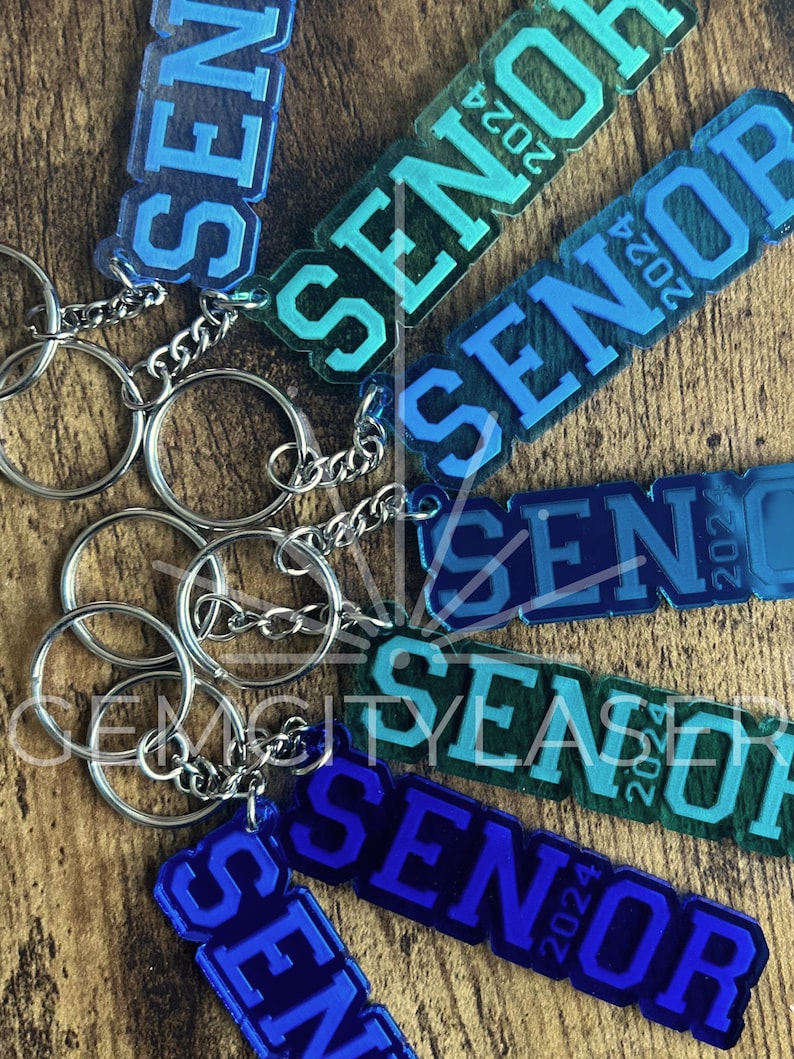 Pu&ograve; includere: Un set di sei portachiavi con la parola "Senior" e l'anno "2024" su ciascuno. I portachiavi sono realizzati in acrilico trasparente con un gradiente blu, verde o viola. I portachiavi sono attaccati a anelli portachiavi argentati.
