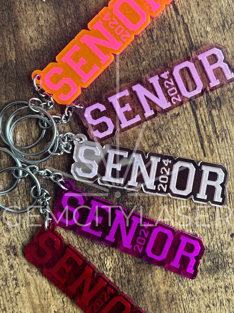 Pu&ograve; includere: Un set di cinque portachiavi con la parola "Senior" e l'anno "2024" su di essi. I portachiavi sono realizzati in acrilico trasparente e sono disponibili in una variet&agrave; di colori, tra cui rosa, arancione, viola e rosso.