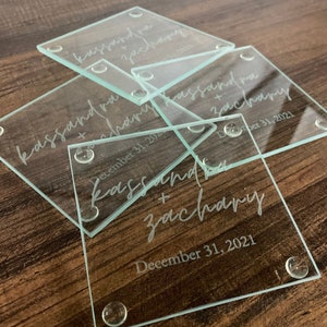 May include: Clear glass square coasters with rounded corners. Each coaster has the names "kassandra + zachary" and the date "December 31, 2021" etched on the surface. Small, clear, rounded feet are on each corner.