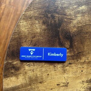 以下が含まれることがあります： 「Kimberly」というテキストと「Erdey Searcy Eye Group, Vision for Life」のロゴが書かれた青い名札