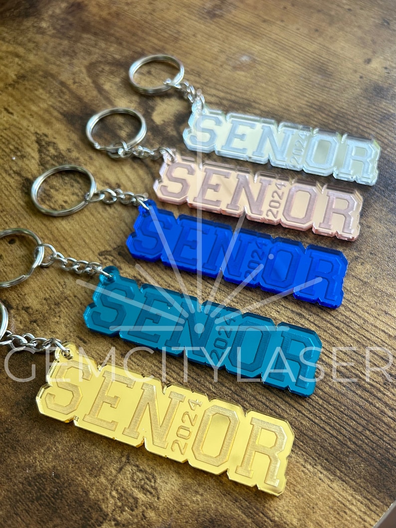 Pu&ograve; includere: Cinque portachiavi con la scritta "Senior 2024" in diversi colori. I portachiavi sono realizzati in acrilico e hanno una finitura a specchio. I colori sono argento, oro rosa, blu, blu verde e oro.