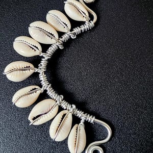 May include: A silver-coloured ear cuff adorned with seven cream-coloured cowrie shells. Each shell is individually wrapped in wire, which is intricately woven along a curved design. A decorative spiral is at the end.