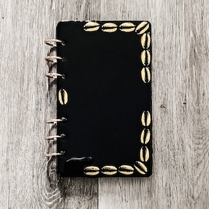 May include: Black notebook with gold shell accents around the edges. The notebook has a ring binder system.