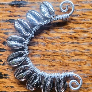 May include: A silver-toned ear cuff featuring a curved wire frame adorned with nine metallic cowrie shells. The wire is wrapped around each shell and terminates in two small, circular spirals at both ends of the crescent-shaped structure.