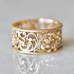 Può includere: Un anello color oro con un intricato design traforato. L'anello presenta un motivo ripetuto di motivi floreali e a foglie vorticose. La fascia dell'anello è larga circa 1,3 cm, con una superficie strutturata e una finitura lucida.