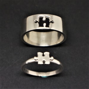 May include: Two silver rings featuring a puzzle piece design. One is a wide band, the other a thinner band. The puzzle piece is cut out, creating a negative space. The rings are shown on a black surface.