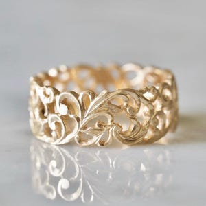 Pode incluir: Um anel dourado com um design intrincado e vazado. O anel apresenta um padrão repetitivo de folhas em espiral e elementos florais. A superfície do anel reflete a luz, criando um efeito brilhante. O anel é provavelmente feito de ouro ou metal dourado.