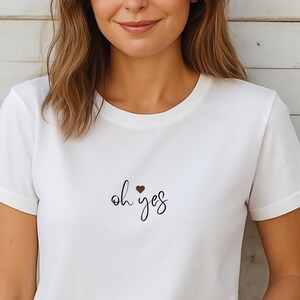 Pode incluir: Camiseta branca com a frase "oh yes" escrita à mão, com um pequeno coração vermelho acima da palavra "yes". A camiseta tem mangas curtas e gola redonda.