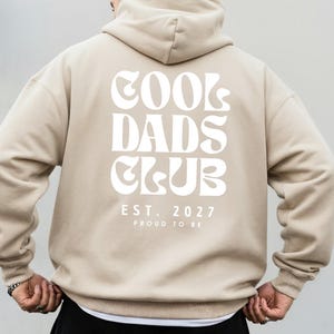 Könnte beinhalten: Ein beigefarbener Kapuzenpullover mit weißem Text auf der Rückseite, der COOL DADS CLUB, EST. 2027 und PROUD TO BE lautet. Das Kleidungsstück hat lange Ärmel, eine Kapuze und einen gerippten Saum und ist von hinten vor einem neutralen Hintergrund zu sehen.