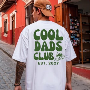 Könnte beinhalten: Ein weißes T-Shirt mit kurzen Ärmeln und dunkelgrünem Aufdruck auf der Rückseite mit der Aufschrift COOL DADS CLUB, einer kleinen Grafik einer Schüssel auf einem Skateboard und dem Text EST. 2027 darunter. Dazu wird eine beige Baseballkappe getragen.