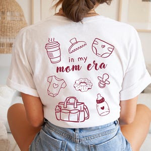 Könnte beinhalten: Ein weißes Kurzarm-T-Shirt mit einer kastanienbraunen Grafik auf der Rückseite. Das Design enthält den Text 'in my mom era', umgeben von Illustrationen eines Kaffeebechers, einer Haarspange, einer Windel, eines Babybodys, eines Haargummis, eines Schnullers, einer Wickeltasche und einer Babyflasche.