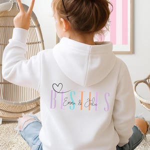 以下が含まれることがあります： 背中にパステルカラーの「BESTIES」の文字と「Emma & Julie」の名前がプリントされた白いパーカー。黒いハートが「BESTIES」の上にあります。パーカーが着用されています。