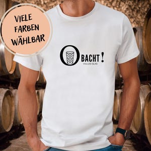 May include: White t-shirt with a black graphic of a glass and the word "BACHT!" in black. The shirt also has the words "VOLLES GLAS" and "VIELE FARBEN WÄHLBAR". The shirt is being worn in front of a background of wooden barrels.