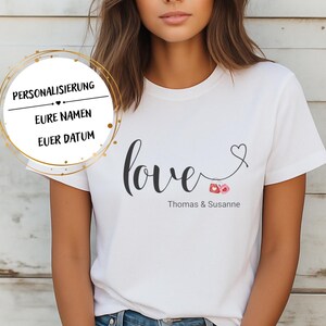 Peut inclure: T-shirt blanc avec le mot "love" en cursive, un cœur et les noms "Thomas & Susanne" imprimés en noir. Le t-shirt comporte également du texte dans un cercle doré qui dit "PERSONNALISATION, VOS NOMS, VOTRE DATE".