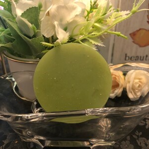 May include: A round, green soap bar with a speckled texture sits in a clear glass bathtub-shaped dish. The dish has three legs and is on a black surface.