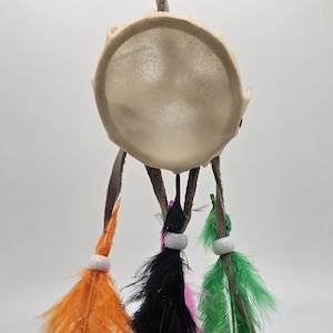 May include: A beige dreamcatcher with three feathers. The feathers are orange, black, and green. Each feather has a white bead at the end.