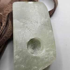 May include: A light green stone pipe with a round hole in the center. The pipe is attached to a brown leather strap.