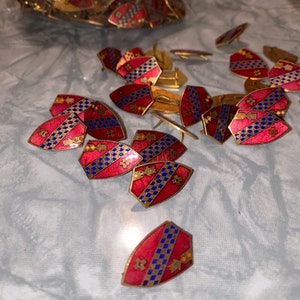 May include: A collection of small, red and blue shield-shaped pins with gold accents. The pins have a checkered pattern and are scattered on a white surface.