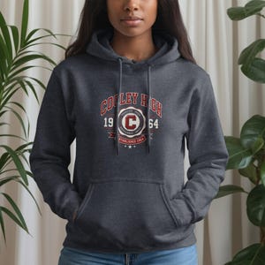 May include: A dark charcoal gray hooded sweatshirt featuring a front kangaroo pocket and matching drawstrings. The chest displays a red and white graphic with the text Cooley High, the numbers 19 and 64, and a central emblem with the letter C.