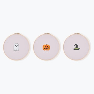 Può includere: Tre cerchi da ricamo a punto croce con disegni a tema Halloween. I disegni includono un fantasma bianco, una zucca arancione e un cappello da strega nero con una fascia verde. Ogni disegno è centrato su un tessuto lilla chiaro.