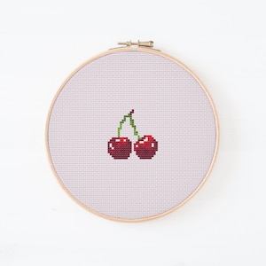 Peut inclure: Broderie au point de croix de deux cerises rouges avec des tiges vertes, sur fond lilas clair, encadrée dans un cercle en bois. Le design simple et coloré est parfait pour la décoration intérieure et ajoute une touche artisanale.
