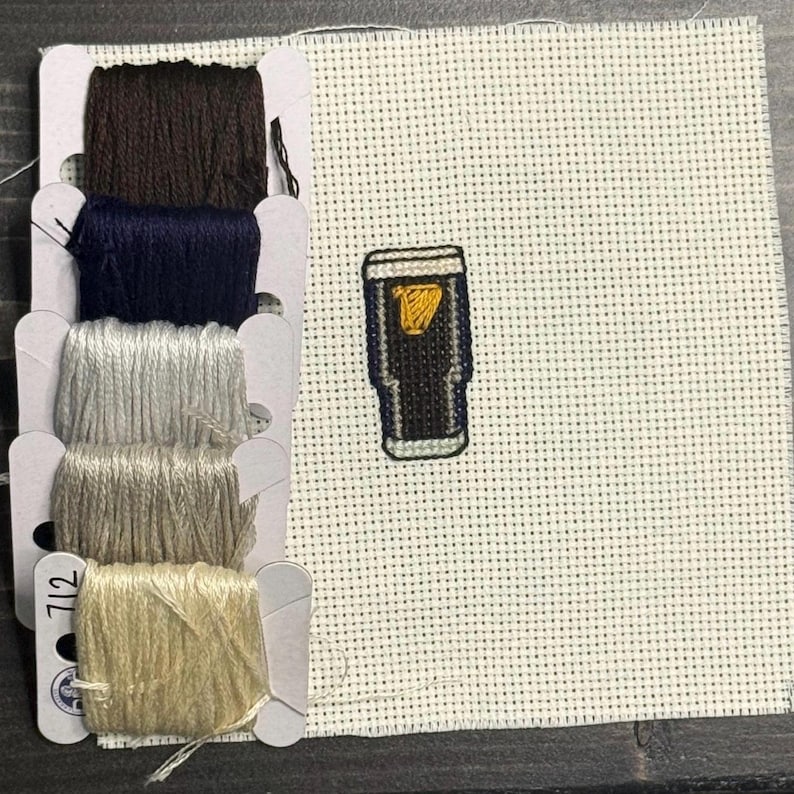May include: A cross-stitch project featuring a pint of stout beer with a gold head. The image includes a selection of embroidery floss in brown, navy, silver, tan, and light yellow colors. The project is on a white dotted canvas.