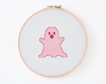 May include: A cross-stitch embroidery hoop featuring a pink ghost design. The ghost has a simple, pixelated appearance with a smiling mouth and small eyes. The embroidery is set against a white fabric background, framed by a wooden hoop.