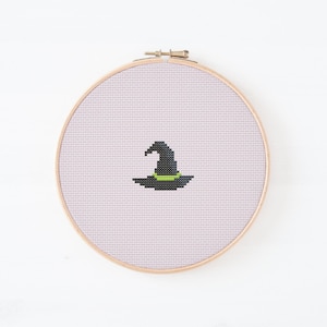 May include: A cross-stitch embroidery hoop featuring a black witch's hat with a green band. The hat is centered on a light purple fabric, set within a wooden embroidery hoop. The design is simple and evokes a Halloween theme.
