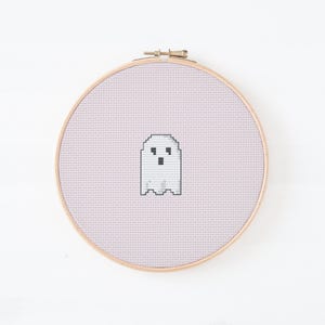 May include: A cross-stitch embroidery hoop featuring a pixelated ghost design. The ghost is white with black eyes and mouth, set against a light pink background. The wooden hoop is round and has a metal clasp.