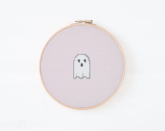 Puede incluir: Un bastidor de bordado de punto de cruz con un diseño de fantasma pixelado. El fantasma es blanco con ojos y boca negros, sobre un fondo rosa claro. El bastidor de madera es redondo y tiene un cierre metálico.