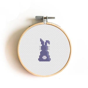 May include: A cross-stitch embroidery hoop featuring a purple bunny design. The bunny is centered on a white fabric background, secured within a wooden hoop. The hoop has a metal tightening mechanism at the top.